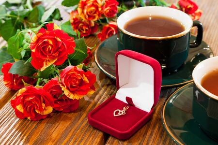 gold jewelry in red velvet box, cups of tea and roses bouquet on wooden tableの写真素材
