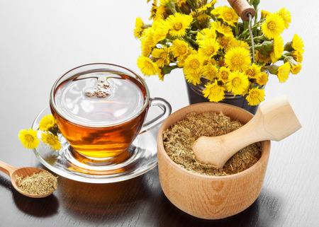 healthy tea in glass mug, bucket with coltsfoot flowers and mortar on table, herbal medicineの写真素材