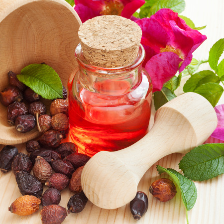 essential oil in glass bottle, dried rose-hip berries in wooden mortar and rose hip flowers on tableの写真素材