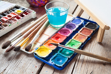 Box of watercolor paints, art brushes, glass of water and easel with canvas or paper on old wooden table.の写真素材