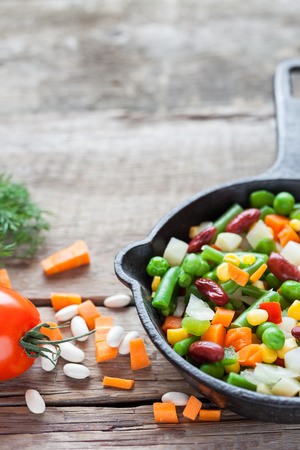 Mixed vegetable meal in old iron frying pan closeup and ingredients on wooden rustic table.の写真素材