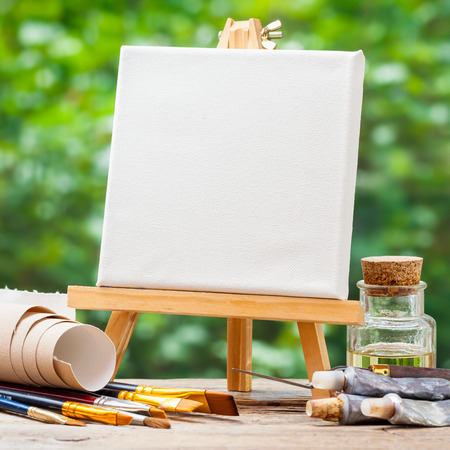 A blank canvas on easel, artistic paintbrushes, tubes of oil paint and bottle of flaxseed oil.