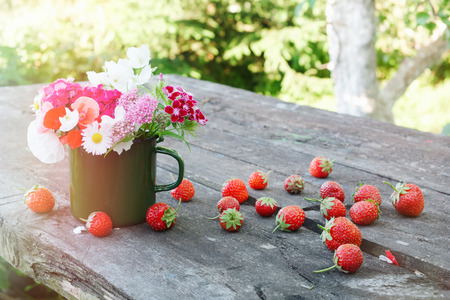 Bouquet of garden flowers in green enameled mug and strawberries on old wooden table outdoors.の写真素材