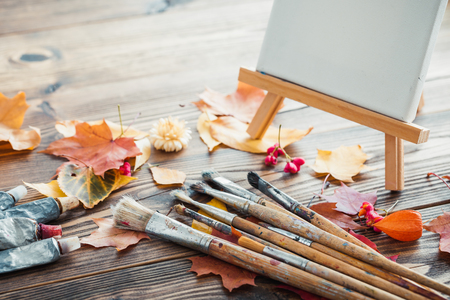 Canvas on easel, paint tubes, brushes for painting and autumn leaves on old wooden background. Retro toned.の写真素材