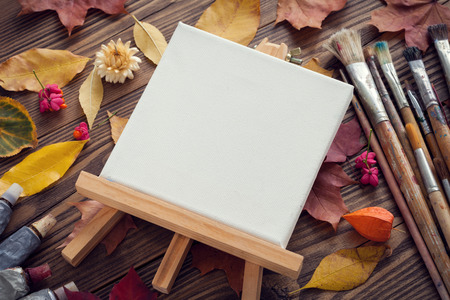 Canvas on easel, paint tubes, brushes for painting and autumn leaves on old wooden desk. Top view. Flat lay. Retro toned.の写真素材