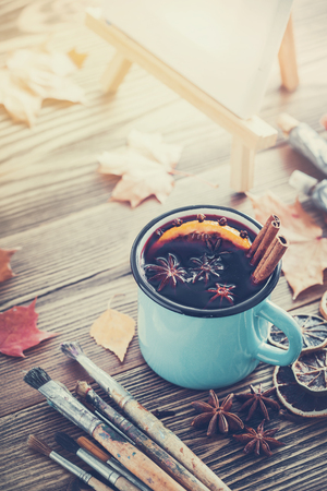 Cup of mulled wine, artist paintbrushes, paints, artistic canvas on easel and autumn leaves on desk.の写真素材