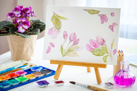 Canvas on easel, watercolor sketch, paints, brushes and violet senpolia flower in pot.の写真素材