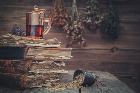 Cup of healthy tea or tincture, mortar of healthy daisy herbs and stack of old books. Hanging bunches of medicinal herbs on background.の写真素材