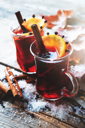 Glass mugs of mulled wine with spices and citrus fruits on wooden table with autumn leaves and snow.の写真素材