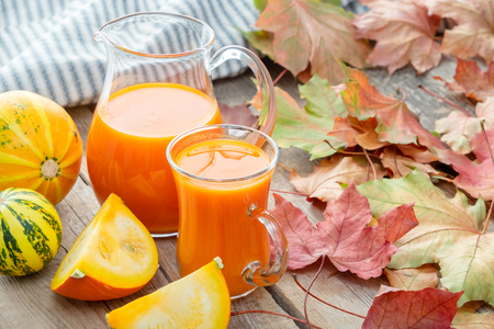 Glass jug and cup of pumpkin juice, pumpkins and fallen maple leaves.の写真素材