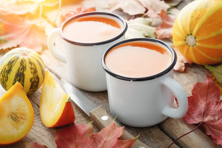 Two white enameled cups of pumpkin juice, pumpkins and fallen maple leaves on table.の写真素材