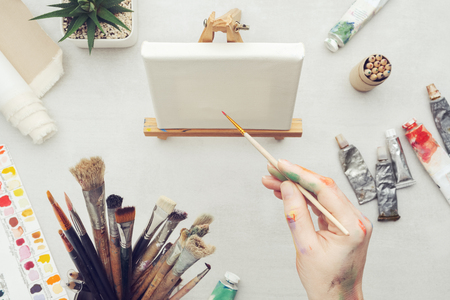 Painter holding a paintbrush in his hand. Canvas on the easel and artistic equipment on desk. Top view.の写真素材