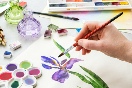 Painter holding a paintbrush in his hand. Watercolor drawing - Iris flower and artistic equipment on desk. Top view. Painter drawing at working place.の写真素材