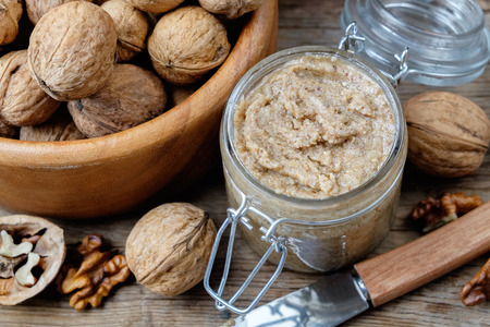 Glass jar of raw organic walnut butter or paste,  fresh nuts in wooden bowl and knife on kitchen table. Top view.の写真素材