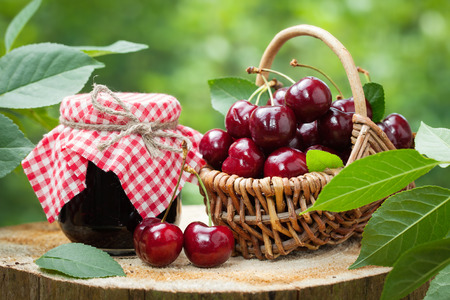 Jar of jam and basket of sweet cherries on background in garden outdoors.の写真素材