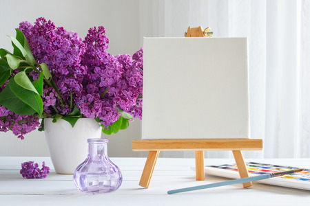 Blank canvas on easel, watercolor paints, brush for painting and lilac flowers on table.の写真素材