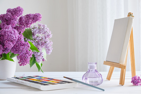 Watercolor paints, canvas on easel and lilac flowers bouquet on white table.の写真素材