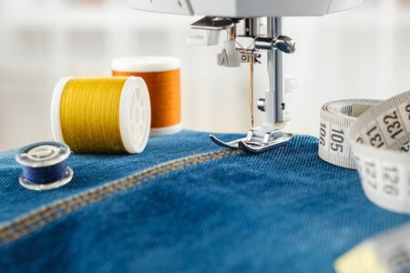 Sewing denim jeans with sewing machine. Close up of needle of sewing machine on the denim fabric stitch, spools of threads and measuring tape.の写真素材