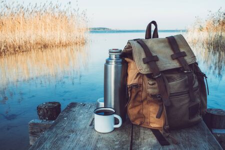Enameled mug of coffee or tea, backpack of traveler on wooden pier on tranquil lake.の写真素材