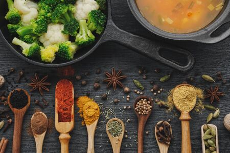 Various aromatic colorful spices and herbs in wooden spoons and scoops. Broccoli and cauliflower on iron frying pan.  Black ceramic saucepan with soup. Top view. の写真素材