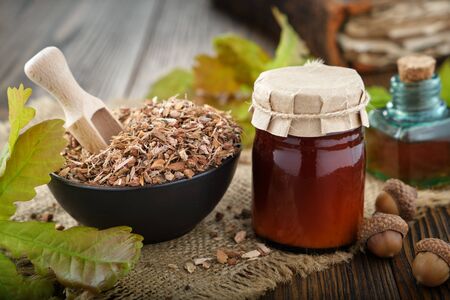 Dry healthy oak bark in black ceramic bowl, jar of salve or balm and green oak leaves and acorns on old bookの写真素材