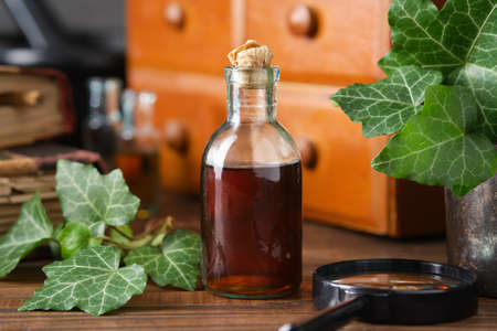 Bottle of ivy leaf syrup or cough tincture. Ivy leaves extract bottle and wooden medicine cabinet with apothecary remedies, old books on background. Alternative herbal medicine.の写真素材