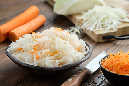 Bowl of sour cabbage, pickled sauerkraut. Fermented cabbage, coleslaw salad. Chopped cabbage on a cutting board and carrots for making sauerkraut on background. Healthy food, diet food.の写真素材