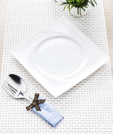 Spoon and fork against empty square white dish on white mat and wooden background の写真素材
