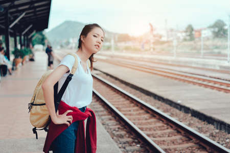 Young girl asian tourist standing with backpack waiting the train going to travel scenery town tour around at train station platform for take the rest, happy and life experience having funの写真素材