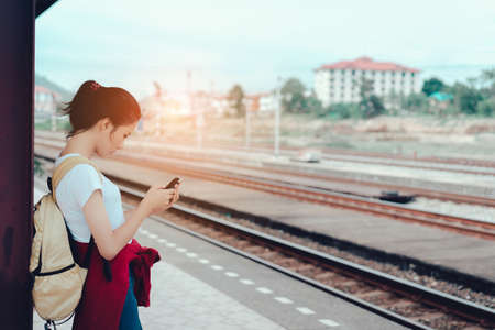 Young girl asian tourist holding smartphone with backpack waiting the train going to travel scenery town tour around at train station platform for take the rest, happy and life experience having funの写真素材