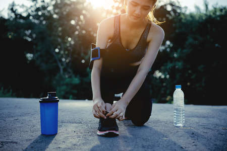 Young girl asian knelt down to do up his shoelaces on street after running exercise with protein shake and water bottleの写真素材