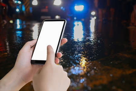 Hand hold mobile blank screen and water on street and color lighting, traffic night illuminated reflection liquid,using the car on the road when it weather rainy season accident, copy space the right.season accident.の写真素材