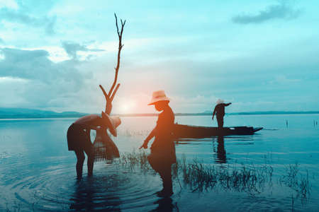 A boy and girl catching fish with a fisherman silhouette on a boat on a river during sunset in Thailand in the background.の写真素材