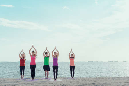 A group of a young men and women practicing yoga on their mat in class exercising on the beach in a modern city during sunrise, relaxing.の写真素材
