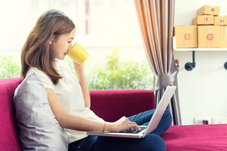 Young asian girl freelancer business private working at home office using laptop computer and drinking coffee with packaging delivery online market on purchase orders to customer.の写真素材