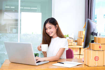 Young asian girl freelancer small business private working at home office with laptop, note, coffee, packaging delivery online market on purchase orders to customer.の写真素材