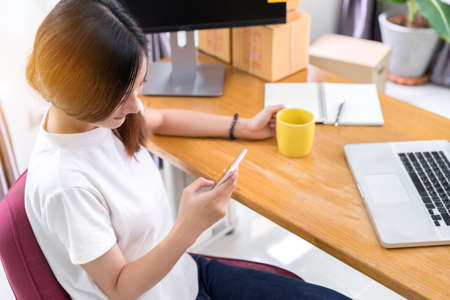 Young asian girl freelancer business private working at home office using phone holding cofee glass with laptop, note, packaging delivery online market on purchase orders to customer.の写真素材