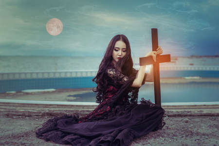 Witch or vampire beautiful woman sitting and holding cross the ghost with  screaming darkness and nightmare, horror of scary fear on hell in halloween festival, and blood moon refurbished by NASAの写真素材