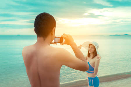 Young man taking photo girlfriend bikini swimsuit and hat with compact camera in beach seaside, Couple honeymoon in holiday and vacation relaxing happiness summer season weekend.の写真素材