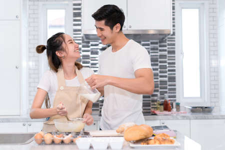 Young asian man and woman couple together making bakery cake and bread with egg, drop milk in the flour happy ralaxing in kitchen at home weekend, cooking lover concept.の写真素材
