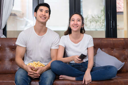 Young asian man and woman couple together lover holding remote watching television screen sitting...
