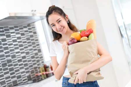 Young asian beautiful girl hold shopping paper bag with vegetables, fruits and bread for making cook...