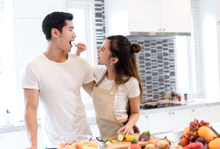 Young asian woman holding vegetables to man eating each for cooking dinner healthy food menu in kitchen at home couple together romanticの写真素材