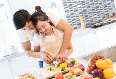 Young Asian man and woman couple together cutting slice vegetables making each salad and food for dinner menu with fruits in kitchen room and home romantic indoor sweet, happyの写真素材