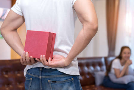 Young asian couple together, man hide holding surprise box heart gift to woman on sofa in home at living room relax and resting happy lover romantic sweet interior indoor concept.の写真素材