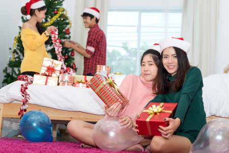 Young Asian group man and women together teenager friend with santa hat holding gift box surprise christmas holidays and  christmas tree background in room at home relaxing celebrating new yearの写真素材