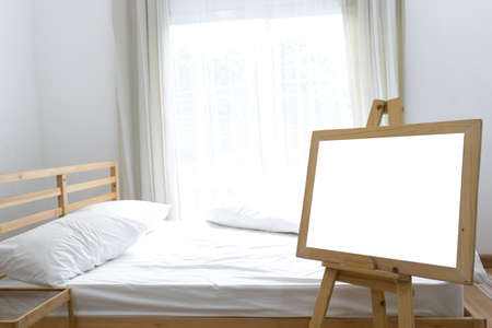 Blank board white frame on wooden standing for poster in bedroom interior with window and pillow の写真素材