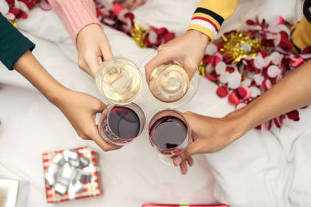 Closeup hands holding glass of wine, cocktail, champagne friend drinking celebration christmas at home party interior on bed and boxの写真素材