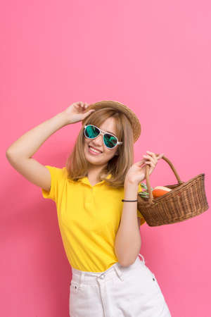 Fashion portrait of girl shopping concet. young beautiful asian with relax dress yellow shirt and white jeans pants, hat, sunglasses, basket vegetables in summer lifestyle on pink background.の写真素材