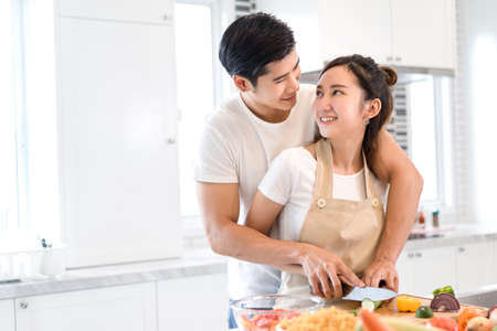 Couple cooking food in kitchen room, Young Asian man and woman together cutting slice vegetables making each salad for dinner menu with fruits at home romantic indoor sweet lover, copy space the left.の写真素材
